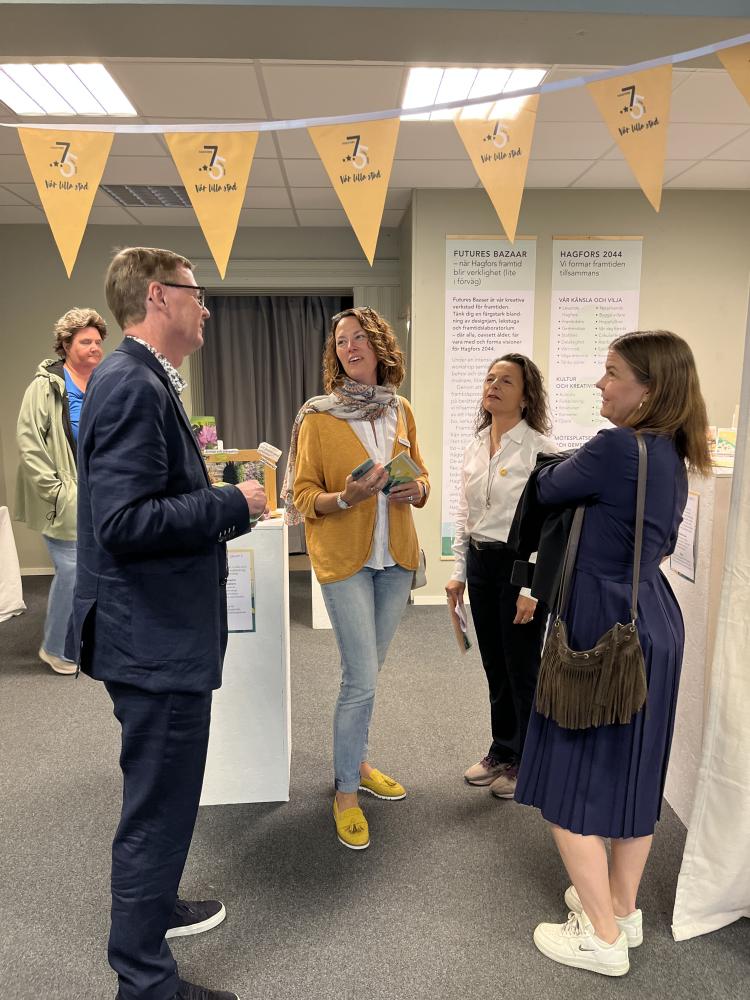 Tre kvinnor och en man pratar med varandra, ståendes i en lokal med grått golv och ljusa väggar. 